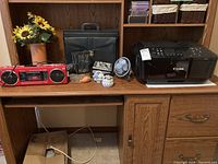 Wide view showing all items on wooden desk including Canon Pixma printer, black briefcase, red candle radio, decorative fan, ceramic jars, picture frame, photo book, and faux sunflower arrangement.