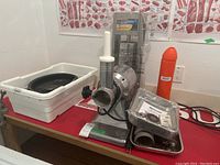 Overall view of meat grinder on stainless steel base next to white plastic tray, vertical sausage stuffer, orange accessory and metal roasting pan