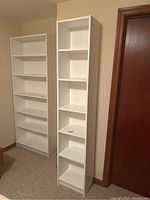 Photo showing two white laminate bookshelves in a beige room: one wider bookshelf with six shelves, and one narrower bookshelf with six shelves.