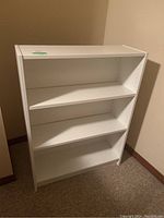 Front angled view of a white laminate bookshelf with three shelves including one angled middle shelf, situated against a wall with light brown baseboard.