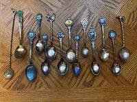 Full set of 12 vintage collector spoons laid out side by side on wooden floor showing varied designs and patina.