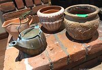 Four pieces shown including three terracotta planters and one brass kettle with coiled handle on a brick surface