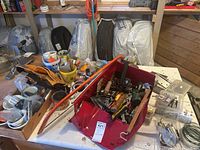 Photo showing a table with a red metal bin full of assorted hand tools, a leather tool belt, and packaged hardware items including screws, nails, and fasteners in the background.
