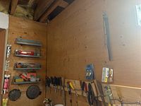 Wide view of the plywood wall where various hand tools, including screwdrivers and pliers, are hanging from hooks, along with some mounted shelves containing saw blades, Kwik Seal product, and other hardware items.