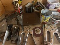 Photo showing multiple metal staple guns, vintage hand plane, round measuring tape, and containers with small hardware such as screws and nails on a wooden benchtop.