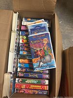 Photo showing an open box with multiple children's VHS tapes stacked inside. Visible titles include The Lion King, The Legend of the North Wind, and others.