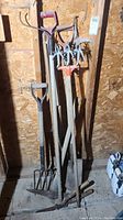Photo showing a variety of yard tools including shovels, hand tillers, pickaxe, and garden shears, all with wooden handles and metal heads. Tools are standing against a wooden wall.