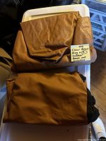 Two brown faux leather bean bag covers folded and laid out on a plastic container, showing tags indicating beans not included.