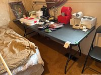 Black plastic folding table with black foldable metal legs, positioned beside a bed and a wall. Table has scratches and wear marks. Various items on the table are not included in the lot.