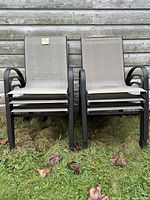 Front view of stack of 6 outdoor garden chairs with black metal frames and beige mesh fabric seats and backs, placed on grass with some leaves around.