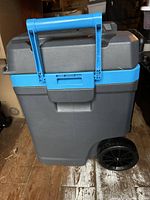 Front angle of the Igloo cooler showing the blue handle, latch, gray body, and one black wheel.
