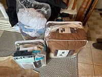 Comforters, blankets, and sheets packaged mostly in plastic cases and bags, displayed on the floor.