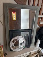 Large rectangular mirror with gray decorative frame leaning against a wall with smaller black framed mirror and round mosaic mirror placed on table in front