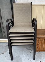 Stacked patio chairs on concrete patio with a wooden cabinet to the right. Chairs have dark metal frames and beige fabric seats.