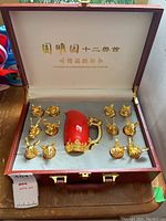 Open maroon case showing 12 gold animal base cups arranged around a red and gold dragon handle pitcher inside gray foam insert.