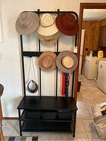 Front view of the black metal shoe rack stand with bench, showing multiple hats hanging on the hooks above the bench with the three bins underneath the bench surface.