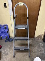 3-step folding stepladder leaning against a door in a garage setting. Visible sturdy metal frame with three black anti-slip steps and a curved top handle. Shows signs of dirt and use.