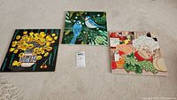 Three ceramic tile art pieces laid out on carpet, showing the full designs of colorful still life themes including flowers, birds, and food.