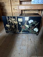 Front view of black lacquer-style trunk with floral and bamboo painted design, showing the locking hardware and decorative feet.