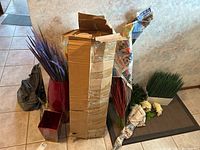 Wide shot showing all items in the lot including large red vase, faux flower stems, white rectangular planter with green faux grass, white artificial roses, small red cube container, and boxed items wrapped in newspaper and cardboard.