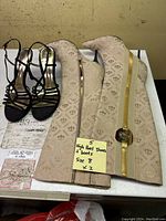 Black strappy high heel shoes and two beige high heel boots with gold zippers arranged on a white surface.
