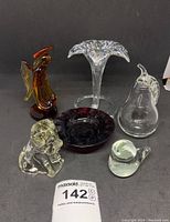 Six glass decorative pieces including amber praying angel, clear glass puppy and pear, amber bird, iridescent flower vase, and dark purple bowl on black background.
