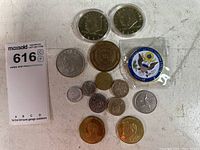 Collection of various coins arranged on a surface with a MaxSold lot tag visible. Includes commemorative and world coins along with medallions.