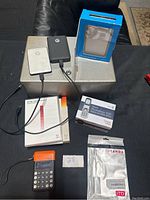 Overview photo showing all items arranged on a black surface including 3 hard drives, boxed phone, calculator, and Adobe software CDs.