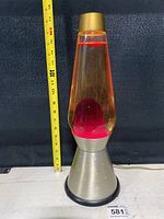 Front view of a 17-inch lava lamp with clear liquid and red wax inside, with a metallic silver base and gold-colored top shown with a tape measure for scale.
