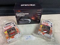 View of SpiEleTek VR-M2 smartphone headset box with two sealed packages of Play Standing Buddy wireless earbuds in front and beside it.