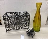 Front and side view of the metal magazine rack with circular design, tall yellow-green glass vase, and black starburst nail sculpture.