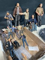 Wide view showing 16 Walking Dead action figures, some standing, others lying in a box, including zombies and main characters with weapons.