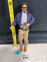 Walter White action figure standing next to a measuring tape showing height about 12 inches, holding a pistol, dressed in blue jacket, checked shirt, beige pants, brown shoes with hat and sunglasses.