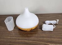 White and wood-tone 300ml aroma diffuser with ribbed design on top, accompanied by clear measuring cup and white power adapter placed on wooden surface.