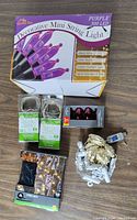Photo showing box of purple decorative mini string lights with 300 LEDs, two packs of 20 clear bulb black wire string lights, pack of 3 red C9 bulbs, coiled gold LED wire lights, and string of warm white decorative bulb-style lights.