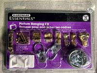 Front of sealed plastic package showing 71 pieces of picture hanging hardware including hooks, nails, wall anchors, screws, and wire with purple and black Hardware Essentials branding and bilingual labels.