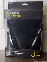 Front view of the retail packaging of J+- Premium stereo headphones. Shows black box with the headphones visible through clear plastic, branding and product features.
