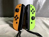 Photo shows a left neon orange Nintendo Switch Joy-Con and a right neon green Nintendo Switch Joy-Con standing upright with attached wrist straps visible.
