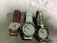 Photo showing three wrist watches on fabric surface. From left to right: rose gold tone metal expandable band watch with white dial, white leather strap watch with large black Arabic numerals, two-tone metal expandable band watch with white dial and gold markers.