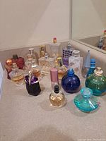 View of twenty partial perfume bottles arranged on a bathroom counter, showing various designs, colors, and partial fillings. Some bottles missing caps or lids.