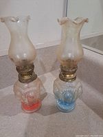 Two small vintage kerosene lamps, one with a red/pink tinted glass base and the other with a blue tinted glass base. Both have clear ruffled glass chimneys and brass wick adjustment knobs.