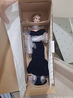 Full doll standing inside original box with arms, legs, and head wrapped in bubble wrap, wearing dark blue velvet off-shoulder gown and a white collar around neck.