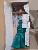 Doll seated in original box, dressed in turquoise satin gown with protective bubble wrap around torso and arms.