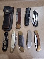 Photo showing eight assorted pocket knives and one black leather sheath arranged on countertop. Handles vary in material and color including wood, black plastic, white/bone-like, and metal.