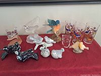 Bird collection displayed on red surface comprising multiple bird figurines made from marble, glass, and ceramic, and three decorated drinking glasses.