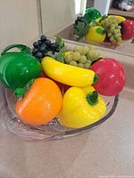 Clear glass bowl filled with colorful blown glass fruit: green apple, red apples, yellow and orange bell peppers, bunches of green and purple grapes, yellow banana.