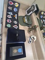 Displaying all items including hat, patches, medal in case, bracelet, t-shirt, and two action figures on a countertop with mirror reflection.