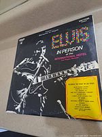 Front cover of "Elvis In Person at the International Hotel" vinyl record album showing Elvis Presley playing guitar with colorful title text and yellow sticker listing songs.