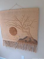 Full view of rectangular handmade wall weaving with natural fibers in beige tone, showing a textured setting sun, tree branches, and a bottom fringe