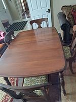 Overhead view of the wooden dining table and five chairs arranged around it. Four chairs share similar carved back designs and upholstery. One chair at the end has different wood and upholstery fabric.